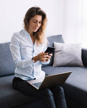 Gömlekli ve pantolonlu genç bir kadın telefonda sohbet ediyor. Dizlerinin üstüne çöküp koltukta oturuyor. Modern dairenin oturma odasında.