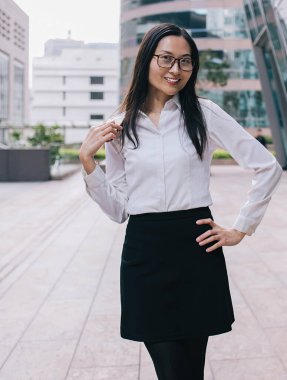 Resmi giyinmiş, gözlüklü, iş merkezinin önünde kameraya bakarak poz veren neşeli, çekici bir Asyalı kadın işçi.