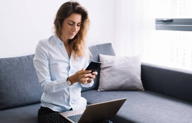 Gömlekli ve pantolonlu genç bir kadın telefonda sohbet ediyor. Dizlerinin üstüne çöküp koltukta oturuyor. Modern dairenin oturma odasında.