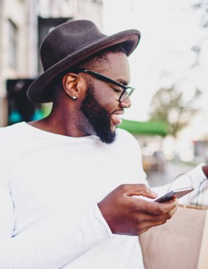 Gülümseyen koyu tenli hippi, alışveriş çantalarına bakıyor. Akıllı telefondan alınmış siparişleri kontrol ediyor. Afro-Amerikan erkeği internetten indirimli alım yapmaktan memnun.