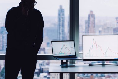 Masaüstünde cihazlarla çalışan çağdaş bir adam ve şehir arka planına sahip pencerenin önünde duran akıllı telefondan çağrı almak