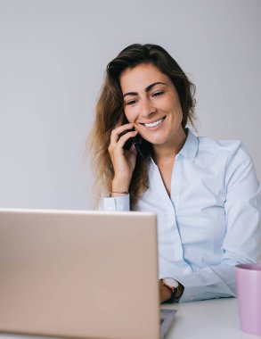 Gömlekli genç esmer, elinde fincanla beyaz masada oturuyor ve telefonda konuşurken gülümsüyor ve bilgisayarında ofisteki bilgisayarında daktilo yazıyor.