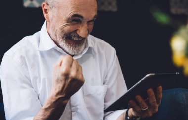 Beyaz tişörtlü, beyaz sakallı mutlu kel adam oturma odasında otururken yumruğunu kaldırmış tablet ekranına bakıyor.