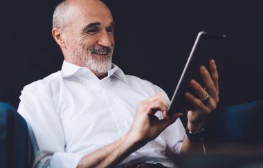 Beyaz tişörtlü, gri sakallı neşeli kel adam oturma odasında otururken tablet ekrana bakıyor.
