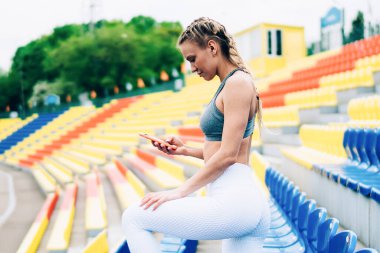 Spor kıyafetli atletik sarışın kadın boş stadyumun sol bacağı üzerinde duruyor ve akıllı telefondan mesajlaşıyor.