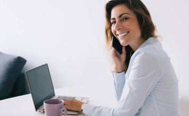 Bardağın ve dizüstü bilgisayarın yanında oturan genç esmerin yan görüntüsü. Beyaz masa üzerinde boş bir boşluk ve telefonla konuşurken gülümseyen ve kameraya bakan.