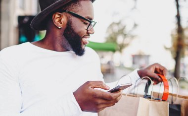 Gülümseyen koyu tenli hippi, alışveriş çantalarına bakıyor. Akıllı telefondan alınmış siparişleri kontrol ediyor. Afro-Amerikan erkeği internetten indirimli alım yapmaktan memnun.