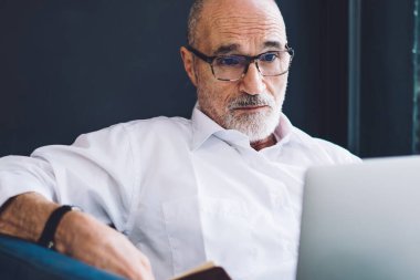 Beyaz tişörtlü, siyah pantolonlu, dizüstü bilgisayarlı yaşlı, sakallı adam oturma odasında pencerenin yanında oturuyor.