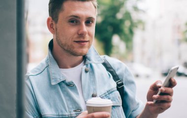 Kot ceketli, tek kullanımlık kahve içen ve sokakta duvara yaslanmış cep telefonu kullanan rahat bir adam.