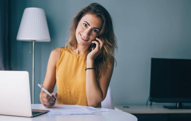 Günlük giysiler içinde gülümseyen genç esmer. Evrakları ve laptopuyla masada oturuyor. Evde kameraya bakıyor ve telefonla konuşuyor.