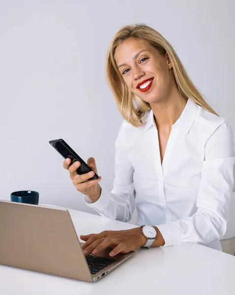 Young female employee with blonde hair sitting at desk with laptop and holding smartphone while smiling and looking at camera in office against white wall