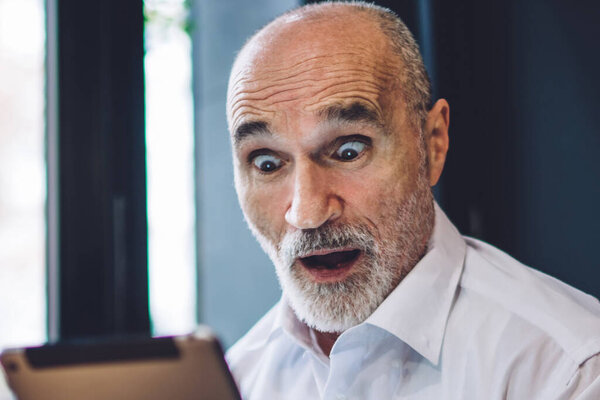 Amazed mature bald male wearing formal clothes using modern tablet with surprise sitting in armchair with open mouth at table at cafe