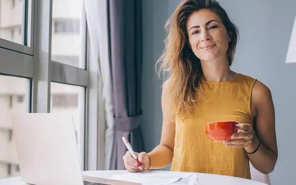 Happy young attractive woman in yellow sleeveless blouse sitting at white round table with laptop and red mug writing on paper at home and looking at camera