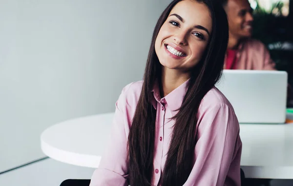 Half length portrait of cheerful woman startup entrepreneur in casual wear sitting in coworking office, positive female employee looking at camera with candid smile on face enjoying working day