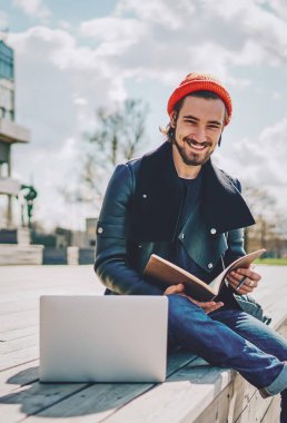 Üniversite sınavına hazırlanmak için vaktini harcayan neşeli beyaz erkek öğrencinin portresi kampüste dizüstü bilgisayar ve eğitim kitabıyla oturuyor, not defteri olan pozitif hipster bir adam.