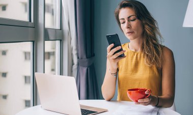 Endişeli ve endişeli kadın serbest çalışan akıllı telefon kullanırken yuvarlak masada oturuyor ve turuncu bardağı iyi aydınlatılmış bir odada tutuyor.