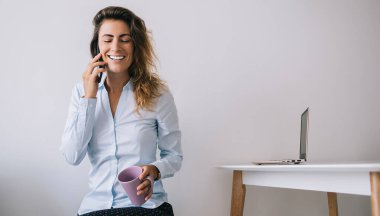 Genç bayan çalışan, elinde kahve fincanı ile gözleri kapalı oturuyor ve telefonda beyaz arka planda konuşurken gülümsüyor.