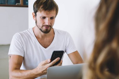 Yetişkin, yakışıklı bir erkek, gündelik giysiler içinde, elinde aletlerle, güzel bir kadının önünde oturmuş kafeteryada akıllı telefon kullanıyor.