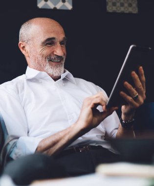 Beyaz tişörtlü, gri sakallı neşeli kel adam oturma odasında otururken tablet ekrana bakıyor.