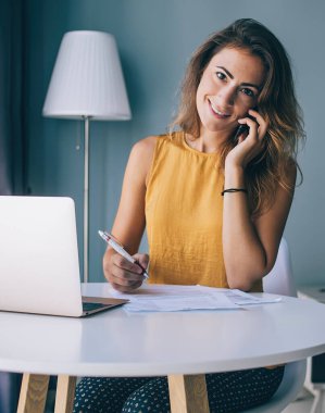 Günlük giysiler içinde gülümseyen genç esmer. Evrakları ve laptopuyla masada oturuyor. Evde kameraya bakıyor ve telefonla konuşuyor.