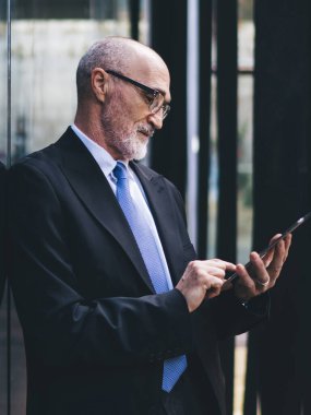 Sokak binasının duvarına yaslanırken, resmi kıyafetli, gözlüklü ve gözlüklü, sakallı, yakışıklı ve kel bir adamın yan görünüşü.