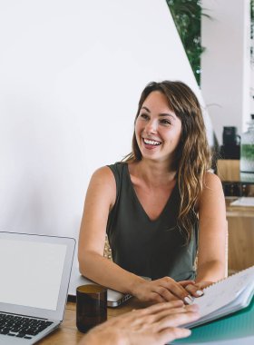 Genç, neşeli, güzel bir kadın. Not defterini beyaz tişörtlü, laptoplu, kafede oturan beyaz sakallı adama gösteriyor.