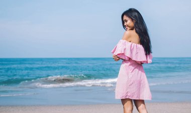 Arkaya bakan pembe elbiseli Endonezyalı genç kadın güneşli bir günde okyanusun arka ucunda gülümsüyor.