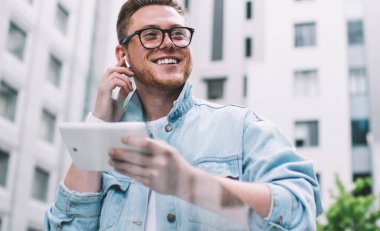 Kot ceketli, kulaklıklı, modern tablet kullanan ve sokakta mutlu bir şekilde gülümseyen mutlu bir adamdan.