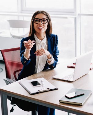 Resmi olarak gülümseyen esmer kadın, ofis içinde masa başında oturmuş iş fikirleri hakkında konuşurken ve işaret ederken profesyonel kadın yöneticinin kırpılmış görüntüsünü takınıyor.