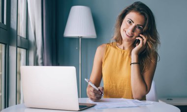 Günlük giysiler içinde gülümseyen genç esmer. Evrakları ve laptopuyla masada oturuyor. Evde kameraya bakıyor ve telefonla konuşuyor.