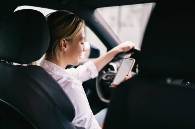 Beyaz tişörtlü, açık renk saçlı bir kadın, siyah bir araçta oturuyor. Direksiyonda oturuyor ve yüzünde gülümseme olan akıllı telefon ekranına bakıyor.