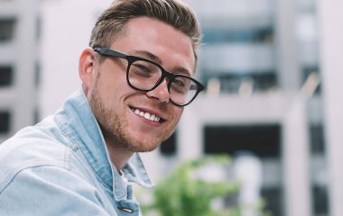 Gözlüklü gülümseyen genç bir adamın yan görüntüsü. Sokakta cam çitlere yaslanmış cep telefonu tutuyor ve kameraya bakıyor.