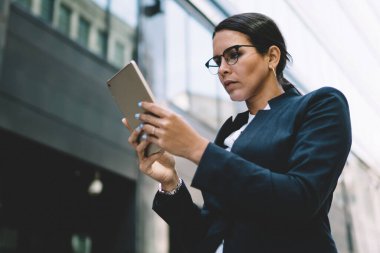 Gözlüklü, siyah saçlı, resmi takım elbiseli, tablet giyen ve şehirde gündüz yürürken ekrana dikkatle bakan bir kadın.