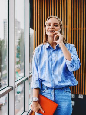 Sıradan giyinmiş neşeli genç bir kadının portresi elinde not defteri tutarken ve akıllı telefon cihazını ararken. Pozitif hipster öğrenci kameraya gülümserken cep telefonuyla konuşuyor.