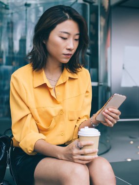 Modern elbiseli düşünceli bir Asyalı kadın sırt çantasıyla oturuyor ve Hong Kong 'da cep telefonuyla sörf yaparken kahve içiyor.