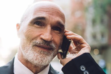 Gülümseyen, takım elbiseli, beyaz sakallı, sokakta yürürken telefonla konuşan, bulanık binalara bakan yaşlı, yakışıklı bir erkek.