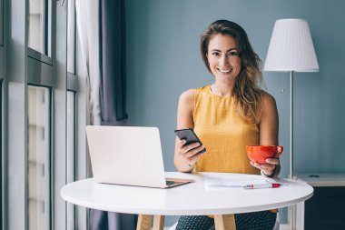 Kameraya bakan mutlu kadın dizüstü bilgisayarla çalışıyor ve sosyal ağları akıllı telefonlarda kullanıyor ve teneffüste bardaktan içiyor.