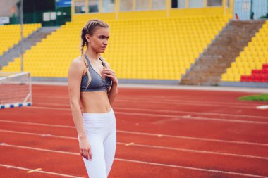 Beyaz taytlı, zayıf bir kadın ve gri bluzlu koşu pistinde duruyor ve teneffüste stadyuma bakıyor.