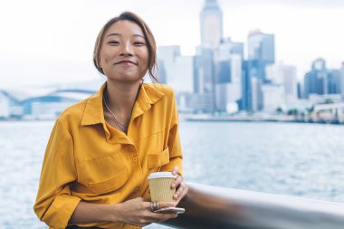 Şık giyinmiş, elinde kahve fincanı ve cep telefonuyla Hong Kong rıhtımına yaslanırken kameraya gülümseyen neşeli genç Asyalı kadın.