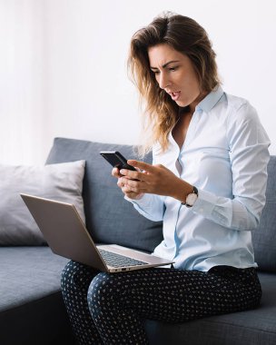 Şaşkın genç kadın cep telefonu kullanıyor koltukta dizlerinin üstüne çöküp oturma odasında çalışıyor.