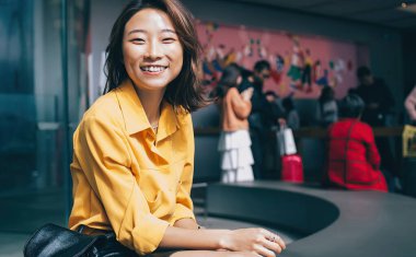 Çekici stil sahibi Asyalı kadın siyah deri etekli ve sırt çantalı, modern koridorda oturmuş Hong Kong kamerasına bakıyor.