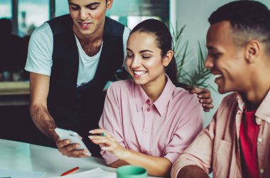 Neşeli erkek ve bayan meslektaşlar sosyal ağlarda cep telefonuyla uygulamayla komik yayınlar okuyor, başlangıçta işbirliğinden sonra, mutlu bir grup insan internet sitesinde şakadan gülümsüyor.