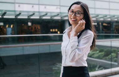 Genç, güzel, neşeli, gözlüklü Asyalı kadın şehir caddesi yakınındaki ofis binasında kameraya bakarak modern akıllı telefondan konuşuyor.