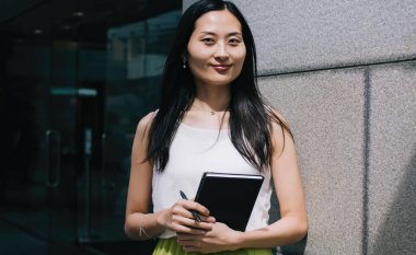 Keyifli stil sahibi genç Asyalı kadın gündüz vakti şehir ofis binasının yanında dururken günlük kıyafetlerle organizatör planlaması yapıyor.