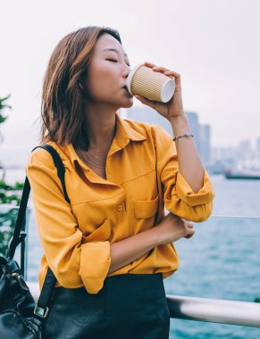 Sarı gömlekli ve sırt çantalı hoş bir Asyalı kadın Hong Kong rıhtımında durmuş kahve içiyor.