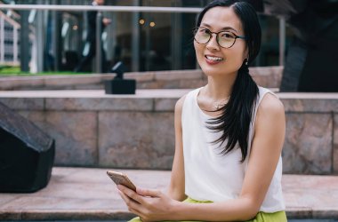 Modern kasaba caddesindeki modern binanın dışında akıllı telefon kullanırken kameraya bakıp gülümseyen gözlüklü etnik bir kadın olması güzel.
