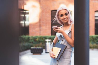 Pembe saçlı, dövmeli, gülümseyen ve kameraya bakan modaya uygun genç bir öğrenci.