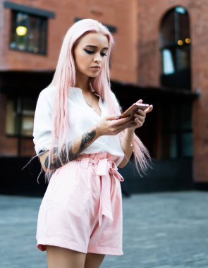 Uzun düz, parlak gül saçlı, yaz kıyafetleri giymiş, caddede beklerken cep telefonuna göz atan genç ve modaya uygun bir kadın.