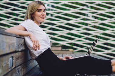 Sıradan kıyafetli genç bir kadının kitap ve dizüstü bilgisayarla ahşap tribünde oturup kameraya bakarken yan görüntüsü.