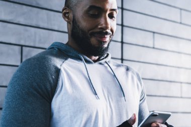 Gri kazak ve eldivenli, spor salonunda gri tuğla duvarın önünde spor yaparken akıllı telefon kullanan gülümseyen yetişkin bir vücut geliştirici.